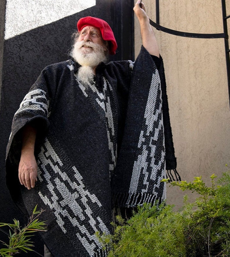 May include: A man with a long white beard and a red beret wears a black and white patterned poncho. He is standing in front of a wall with a black and white geometric design.