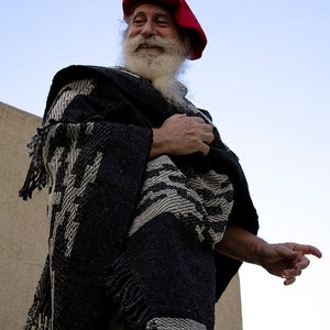 May include: A man with a long white beard wears a red beret and a black and white patterned poncho.