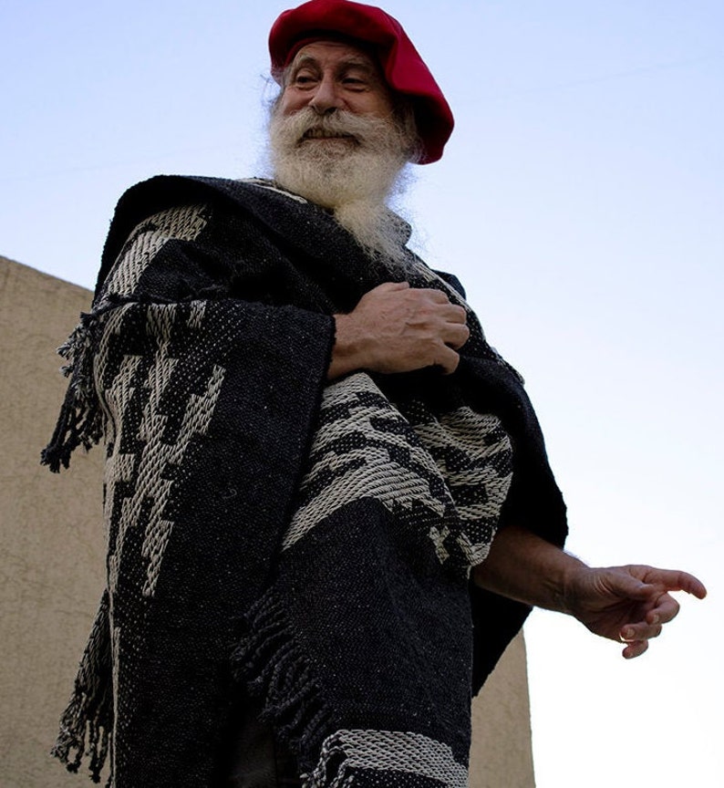 May include: A man with a long white beard wears a red beret and a black and white patterned poncho.