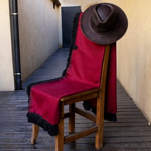 Peut inclure: Une couverture en laine bordeaux avec des franges noires drap&eacute; sur une chaise en bois. Un chapeau de cowboy en cuir marron est pos&eacute; sur la couverture.