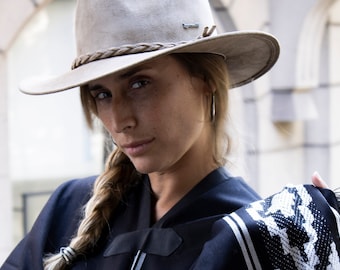 May include: A woman wearing a wide-brimmed, light brown felt hat with a braided band, a black shirt, and a black and white striped shawl. Her hair is braided and pulled to the side.