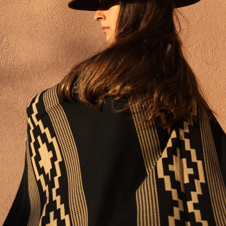 May include: A black and tan striped poncho with a geometric pattern. The poncho is worn by a person with long brown hair and a brown hat.