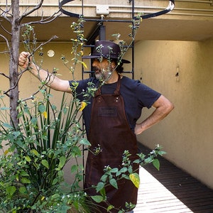 May include: A man wearing a brown leather apron and a black hat stands in a garden. He is looking at a plant with green leaves and small black berries.