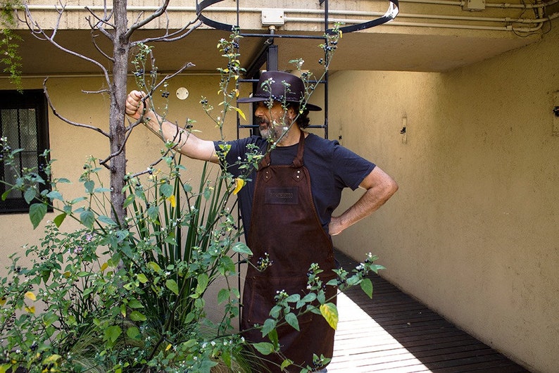 May include: A man wearing a brown leather apron and a black hat stands in a garden. He is looking at a plant with green leaves and small black berries.