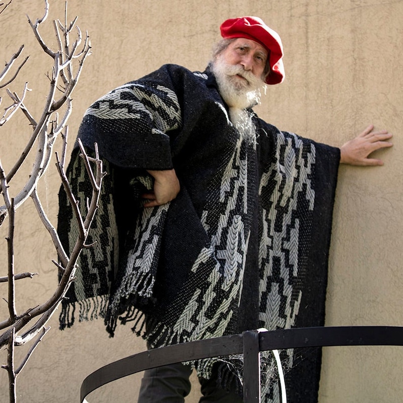 May include: A man wearing a black and white patterned poncho with a red and white beret. He has a long white beard and is standing in front of a tan wall.