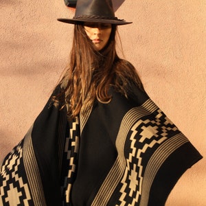 May include: A black and tan patterned poncho with a geometric design. The poncho is worn by a person wearing a brown leather hat.