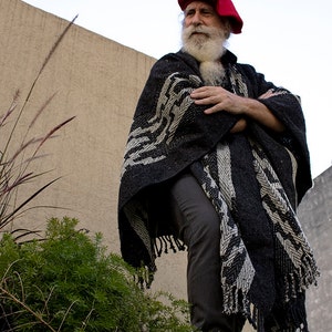 May include: A man with a long white beard and a red beret wears a black and white patterned poncho. He is standing in front of a beige wall.