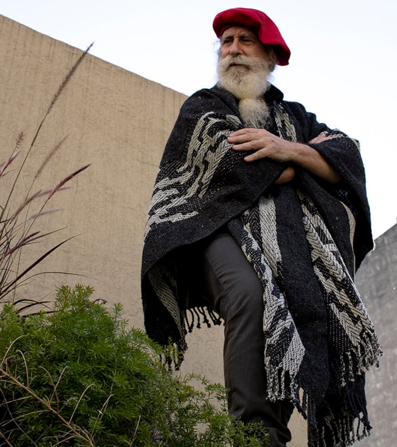 May include: A man with a long white beard and a red beret wears a black and white patterned poncho. He is standing in front of a beige wall.