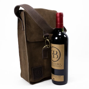 Puede incluir: Un porta botellas de vino de gamuza marrón con una correa de cuero y un cierre de metal. El porta botellas contiene una botella de vino tinto con una etiqueta de papel marrón que dice "Boyero."