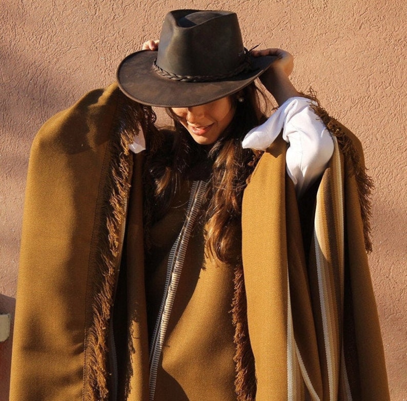 May include: A brown poncho with fringe trim and a white stripe down the side. The poncho is worn by a person wearing a brown leather cowboy hat.