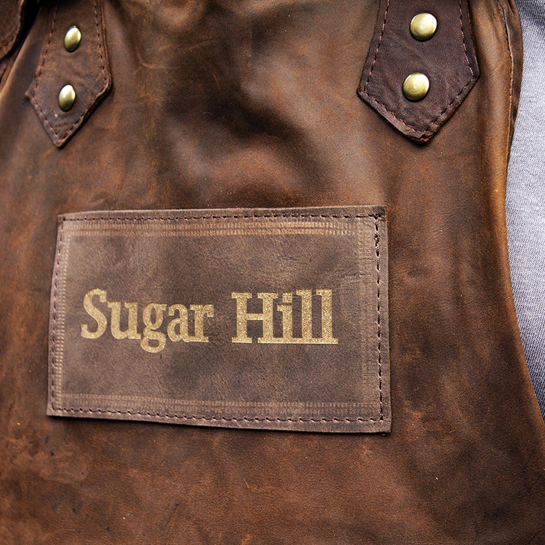 May include: A brown leather apron with a patch that says "Sugar Hill" in gold lettering.