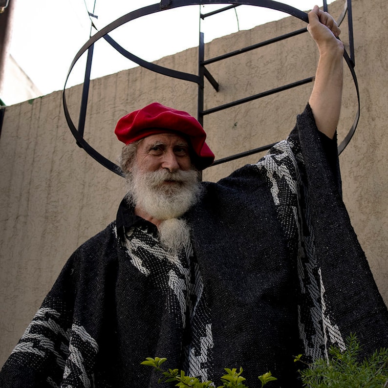 May include: A man with a long white beard and a red beret is wearing a black and white poncho. He is standing in front of a wall with a metal ladder.