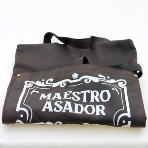 May include: A brown leather apron with a white embroidered design that says "Maestro Asador".