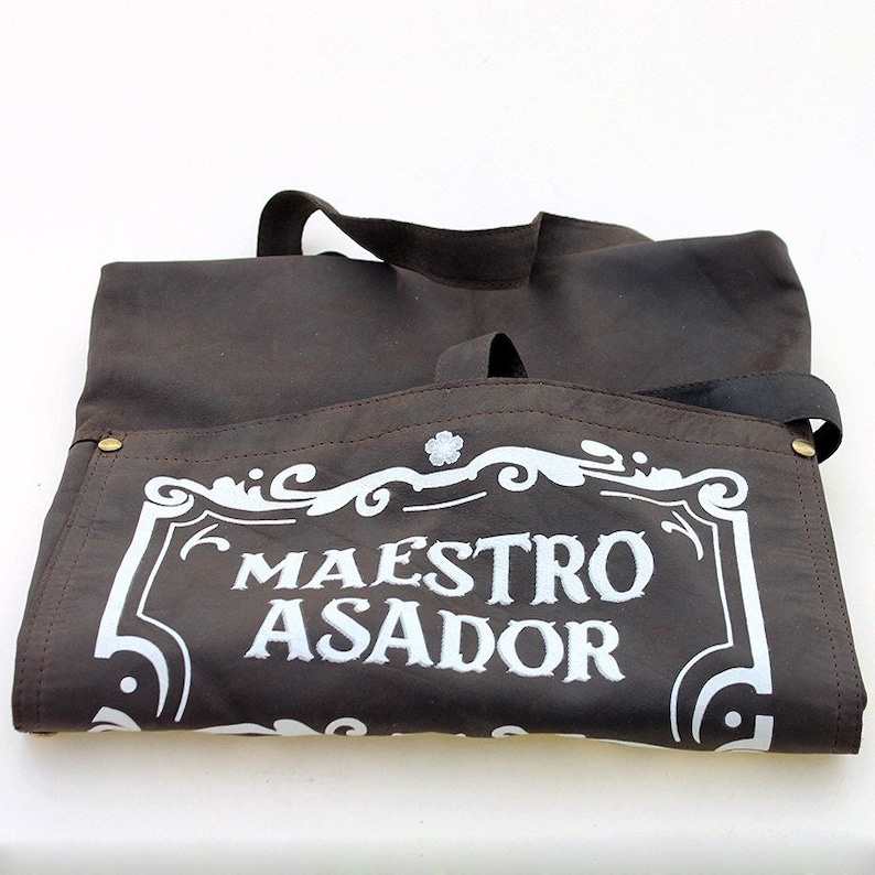May include: A brown leather apron with a white embroidered design that says "Maestro Asador".