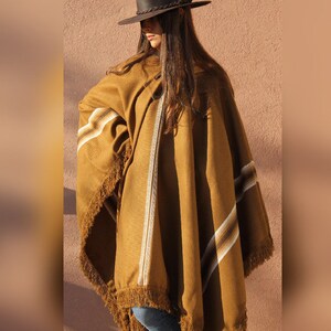 May include: A brown poncho with white and brown stripes and fringe trim. The poncho is worn by a person wearing a brown hat.