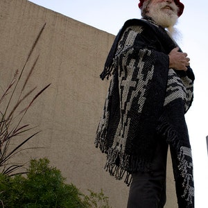 May include: A man wearing a black and white patterned poncho with a red beret and a long white beard. He is standing in front of a tan wall.