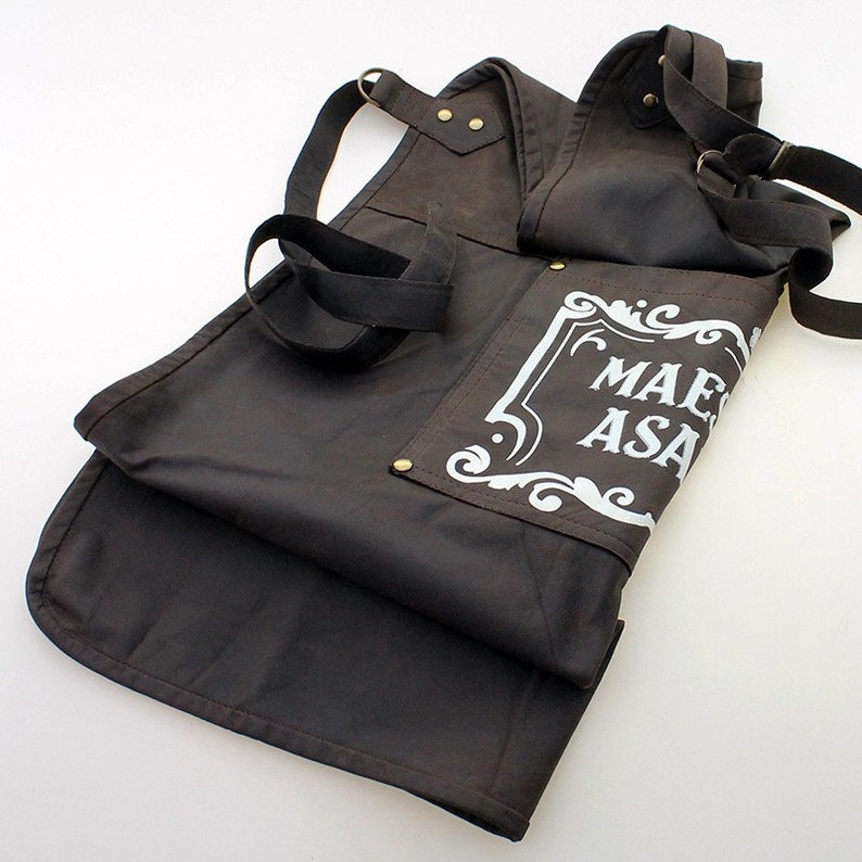 May include: A brown canvas apron with adjustable straps and a white graphic that says "Mae Asa".