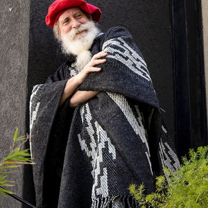 May include: A man with a long white beard and mustache is wearing a red beret and a black and white patterned poncho. He is standing in front of a gray wall.