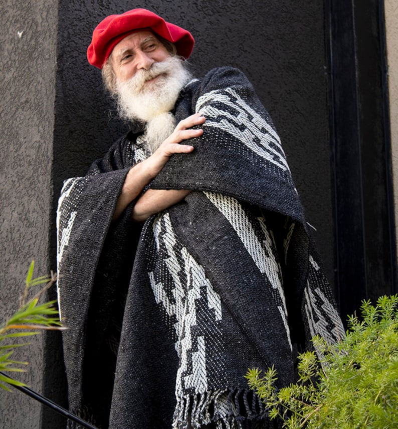 May include: A man with a long white beard and mustache is wearing a red beret and a black and white patterned poncho. He is standing in front of a gray wall.