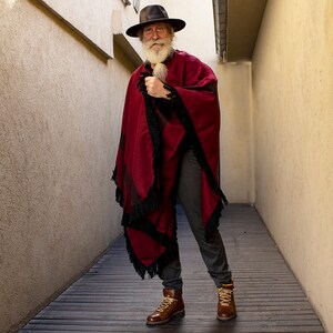 Peut inclure: Un homme portant un poncho rouge et noir &agrave; franges, un chapeau noir et des bottes marron. Il a une longue barbe blanche et se tient dans un couloir &eacute;troit.