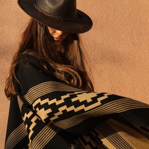 May include: A woman wearing a black and tan patterned poncho with a brown leather cowboy hat.