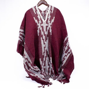 Puede incluir: Un poncho burdeos con un patr&oacute;n geom&eacute;trico blanco. El poncho tiene cuello en V y flecos en el borde inferior. La prenda se exhibe en una percha de madera sobre un fondo blanco.