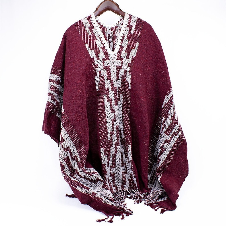 Puede incluir: Un poncho burdeos con un patr&oacute;n geom&eacute;trico blanco. El poncho tiene cuello en V y flecos en el borde inferior. La prenda se exhibe en una percha de madera sobre un fondo blanco.