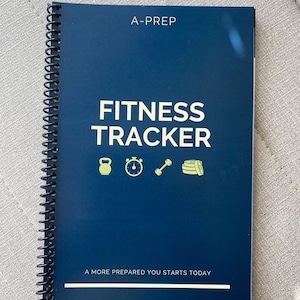 May include: A dark blue spiral-bound fitness tracker notebook with the text A-PREP, FITNESS TRACKER, and A MORE PREPARED YOU STARTS TODAY printed in white. The cover features yellow icons of a kettlebell, stopwatch, dumbbell, and weight plates.