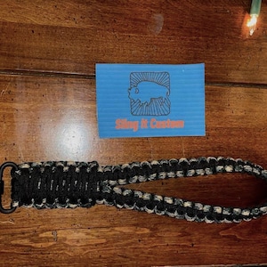 May include: A black and tan paracord sling with a black metal swivel clip. The sling is made with a braided pattern and is approximately 18 inches long. The text "Sling It Custom" is visible on a blue card in the background.