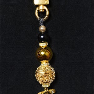 May include: A gold-toned metal keychain featuring a lobster clasp, a small silver bead, a smooth black bead, a large reflective gold bead, and a textured gold sphere encrusted with clear crystals, finished with a small crystal-accented bee charm.