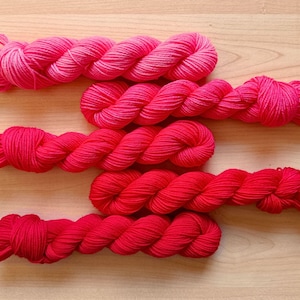 May include: Five skeins of vibrant pink and red yarn are arranged on a light wood surface. The yarn is tightly wound, showcasing a range of shades from light pink to deep red. Ideal for knitting or crochet projects.