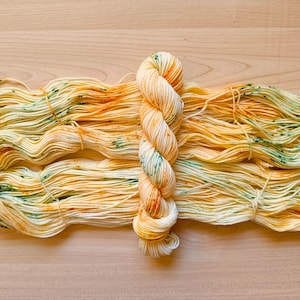 May include: A skein of hand-dyed yarn in shades of yellow, orange, and green. The yarn is twisted into a skein and lies next to a larger amount of loose yarn in the same color palette. The yarn is on a light wood surface.
