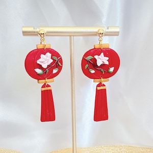 May include: Red lantern earrings with white flower accents and gold detailing. The earrings feature a round shape and a tassel design. They are displayed on a gold stand against a white backdrop.