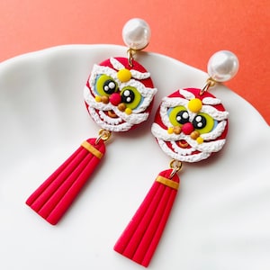May include: A pair of red and white lion dance earrings with gold accents and pearl studs. The earrings feature a round lion head design with a red tassel. The earrings are on a white surface.