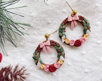 May include: A pair of festive earrings shaped like miniature wreaths. Each wreath features a brown base adorned with green leaves, red berries, and colorful flowers in shades of pink and yellow. A pink bow sits at the top of each earring.