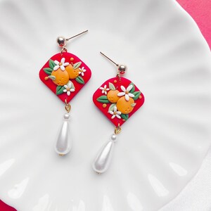 May include: Dangle earrings with a red square design, orange fruit, and white flower details. Each earring has a gold post and a white teardrop pearl. The earrings are displayed on a white plate.