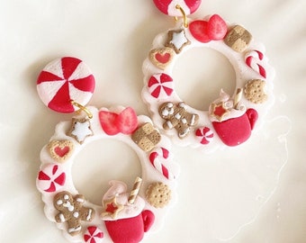 May include: Festive dangle earrings featuring a white wreath design adorned with miniature gingerbread men, candy canes, cookies, and red and white peppermint candies. Each earring has a red and white peppermint candy stud.