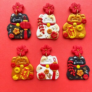 May include: Six pairs of handmade Lucky Cat earrings in various colours, including black, white, and gold. Each earring features a red flower, a waving cat, and decorative elements like lanterns and coins. The earrings are set against a red background.