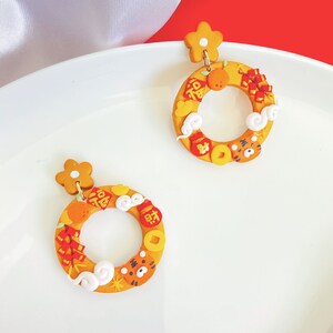 May include: A pair of yellow hoop earrings with a flower-shaped stud. The hoops are adorned with Chinese New Year symbols, including a tiger, coins, clouds, and oranges. The earrings are displayed on a white plate against a red backdrop.