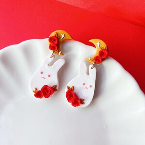May include: A pair of white bunny earrings with red rose detailing and gold crescent moon features. These earrings have a whimsical design, ideal for adding a touch of charm to any outfit.