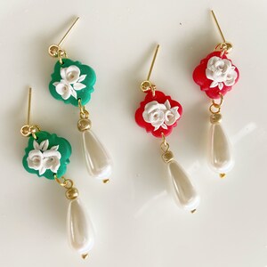 May include: Dangle earrings with a green or red quatrefoil design, adorned with white floral details. Each earring has a gold-coloured post and connecting hardware, with a pearlescent teardrop-shaped bead suspended below. The earrings are shown on a white surface.