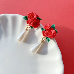 May include: Handmade earrings with red rose clusters and green leaves. Each earring has a gold-coloured hook and a pear-shaped, white faux pearl drop. The earrings are displayed on a white, scalloped plate against a red background.