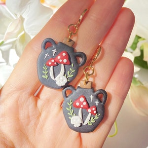 May include: A pair of dark grey cauldron-shaped earrings with gold-coloured hooks. Each earring features red and white mushroom designs, white skulls, and green foliage. The earrings are held in a hand.
