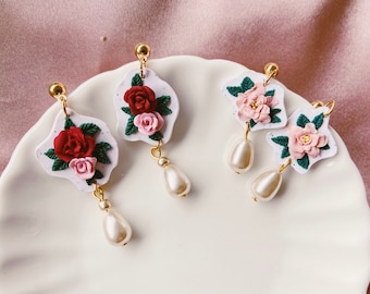 May include: Two pairs of drop earrings featuring white, irregular-shaped backings with sculpted red and pink roses, green leaves, and dangling teardrop-shaped faux pearls attached to gold-toned metal posts and hardware.