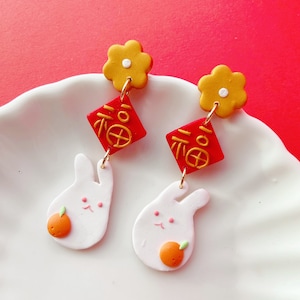 May include: A pair of earrings with a yellow flower, red diamond with gold Chinese characters, and a white bunny with an orange fruit. The earrings are connected with gold-coloured hoops and are set against a red background.