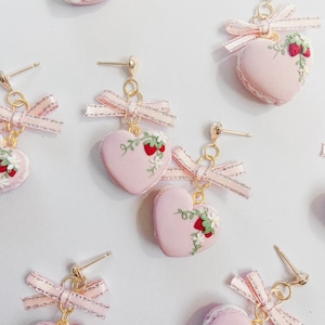 Puede incluir: Pendientes rosas en forma de corazón con diseño de fresas y flores. Los pendientes tienen un poste dorado y están adornados con un lazo de cinta rosa. Los pendientes están diseñados para parecerse a macarons.