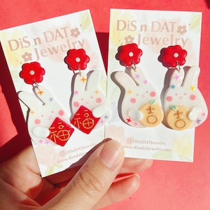May include: A pair of white bunny earrings with red flower accents. The earrings feature a red envelope with gold Chinese characters and a gold-coloured charm. The earrings are on a white card with the text "DiS n DAT Jewelry".