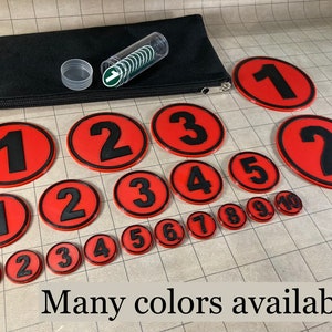 May include: A collection of red and black numbered tokens and a black pouch. The tokens feature black numbers 1-10 on a red background with a black border. A clear tube with green and white tokens is also present. The text "Many colors available!" is at the bottom.