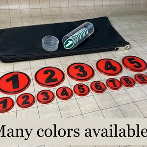 May include: A collection of red and black numbered tokens, ranging from 1 to 10, with a black carrying case and a clear tube containing green and white tokens. The text "Many colors available!" is displayed at the bottom.