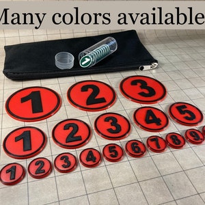 May include: Red and black numbered tokens, ranging from 1 to 10, are displayed. The text "Many colors available!" is at the top. A black pouch and a clear tube containing green and white tokens are also present.